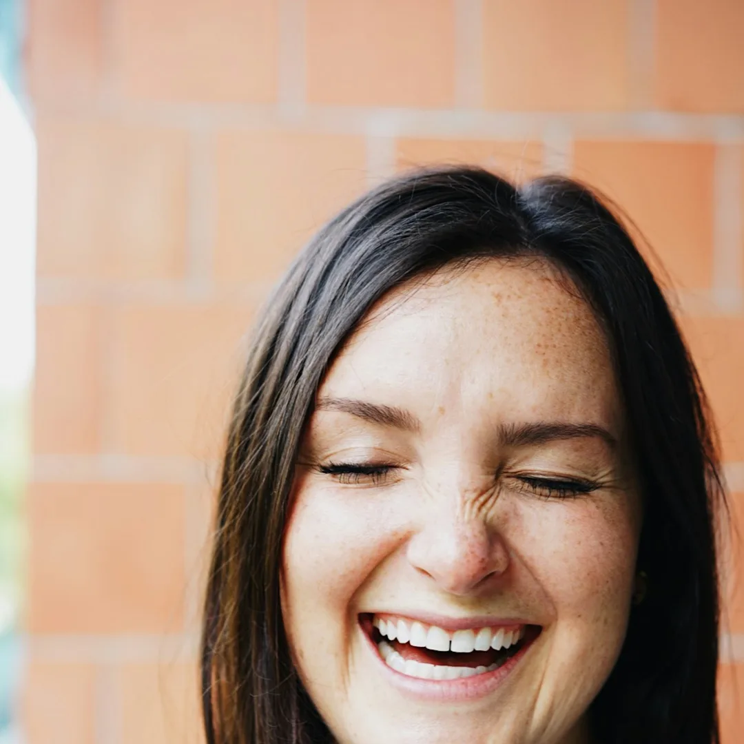 A woman laughing with her eyes closed against a brick wall