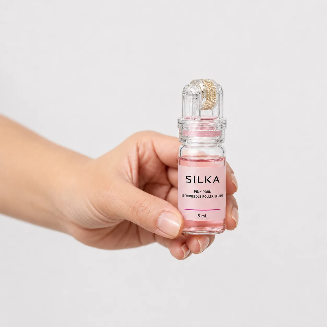 A hand holding the Silka Pink PDRN roller bottle upright, the gold microneedle head under the clear cap
